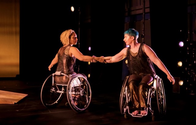 Laurel Lawson and Alice Sheppard dance around each other in a counterbalance turn. They gaze at each other as they hold forearms and extend their opposite arm. Laurel is a white dancer with cropped blue hair and Alice is a multiracial Black woman with short curly hair; they both wear shimmery dark sleeveless costumes. Photo by J. Adam Fenster / University of Rochester